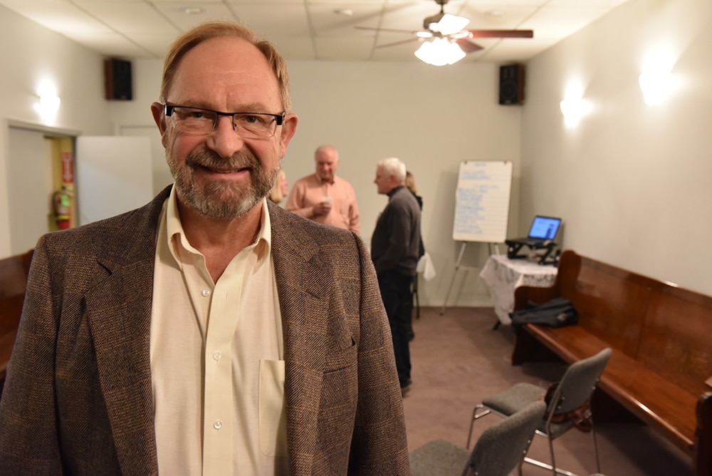 Gary Sulz, an older while man wearing a brown suite and glasses, staring at the camera and smiling. He is standing in a warmly lit conference room with tables and a bench behind him. There are two people in the background talking.