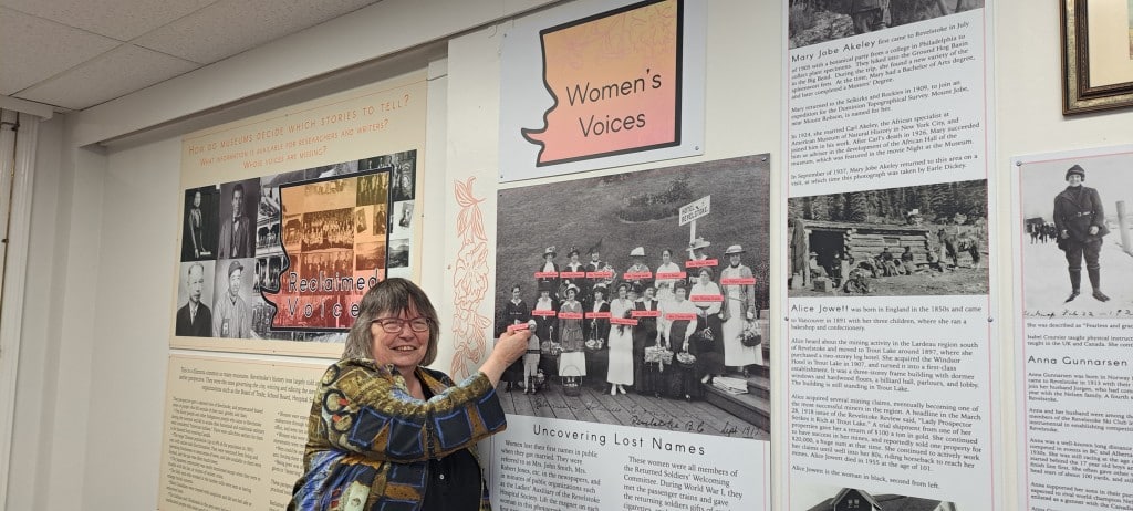 Cathy English, a woman wearing a textile blouse, glasses and smiling widely at the camera, points to names of Revelstoke's early women in Revelstoke Museum and Archives Reclaimed Voices exhibit.