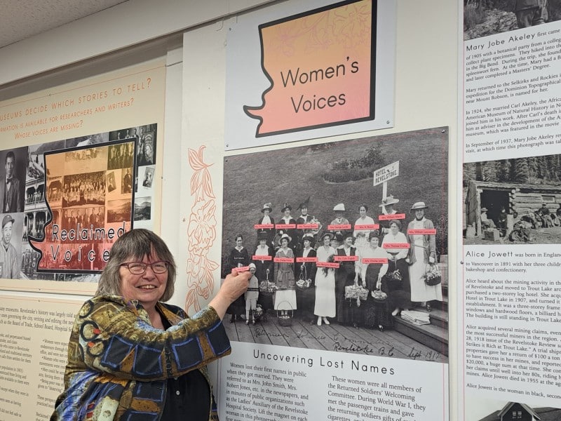 Cathy English, a woman wearing a textile blouse, glasses and smiling widely at the camera, points to names of Revelstoke's early women in Revelstoke Museum and Archives Reclaimed Voices exhibit.