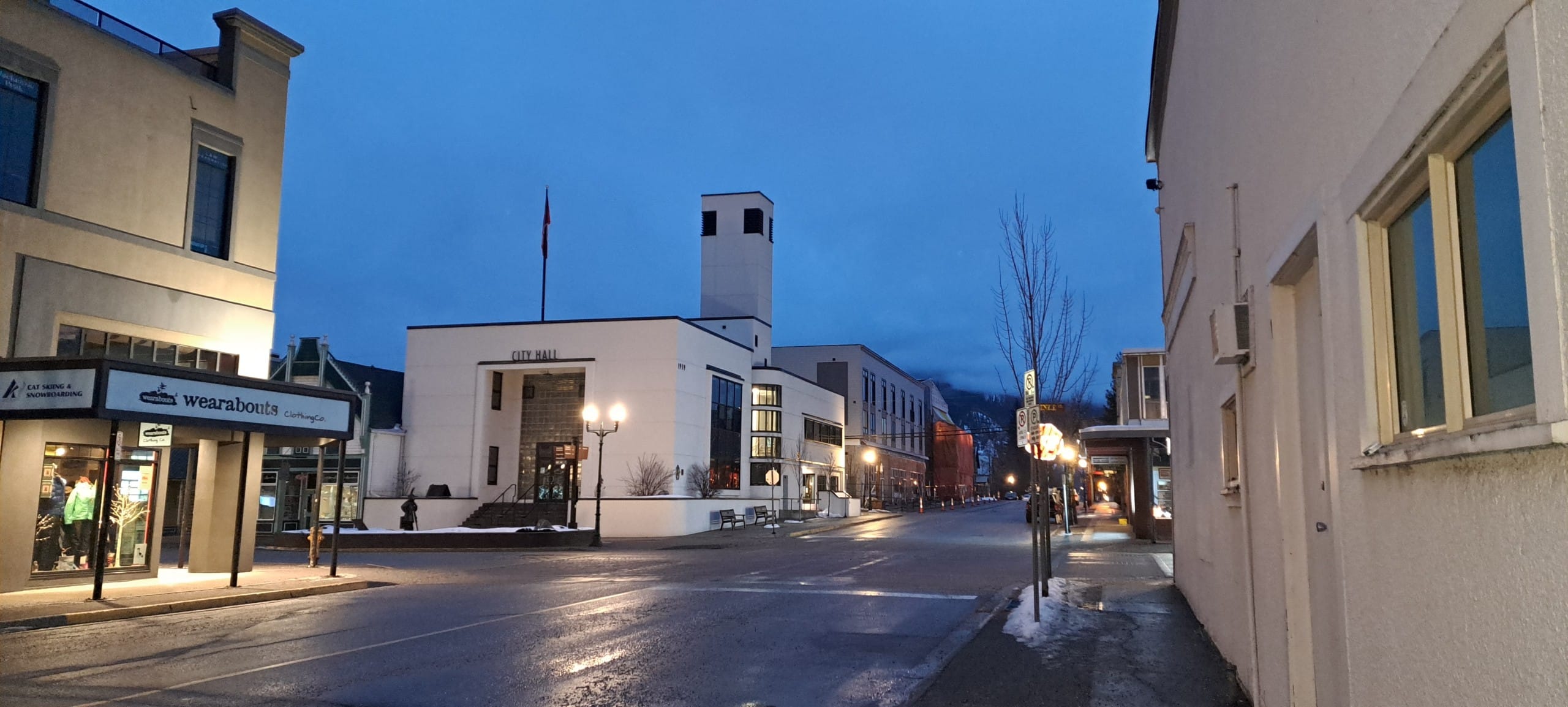 Revelstoke city hall in early night lighting. The building is a large white box shape with two floors. The entrance is sunken into the face of the building.
