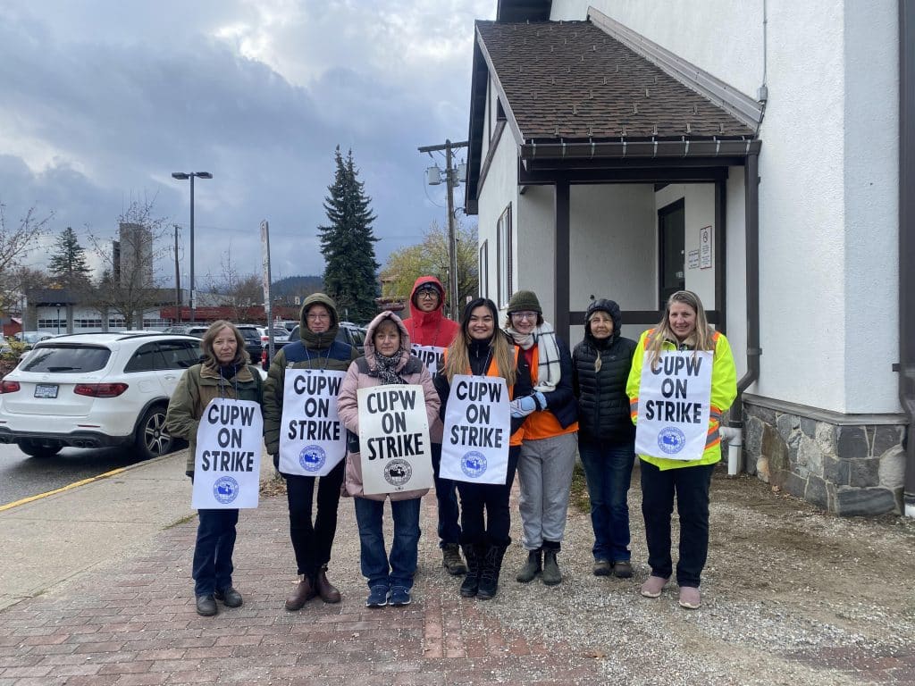 Eight workers stand outside the Canada Post office in Revelstoke, all wearing large, "CUPW on strike" signs.