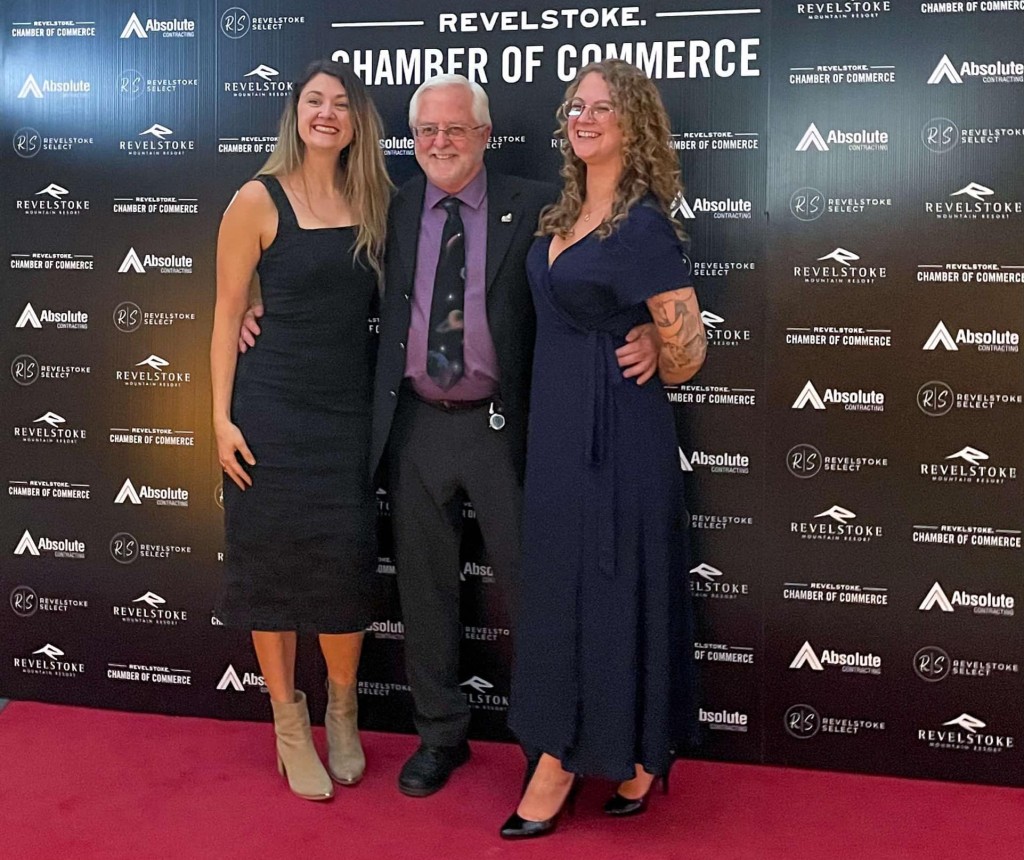 left to right Meghan Porath, a white woman with long brown hair wearing a black cocktail dress, Ken Talbot, an older white man wearing a black suit and purple under shirt and Taylor Sandell, a white woman with curly hai and wearing and dark blue gown dress stand in front of a Revelstoke Business Excellence Awards backdrop as they smile at a camera to the left of the picture.