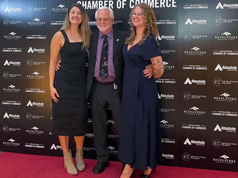 left to right Meghan Porath, a white woman with long brown hair wearing a black cocktail dress, Ken Talbot, an older white man wearing a black suit and purple under shirt and Taylor Sandell, a white woman with curly hai and wearing and dark blue gown dress stand in front of a Revelstoke Business Excellence Awards backdrop as they smile at a camera to the left of the picture.