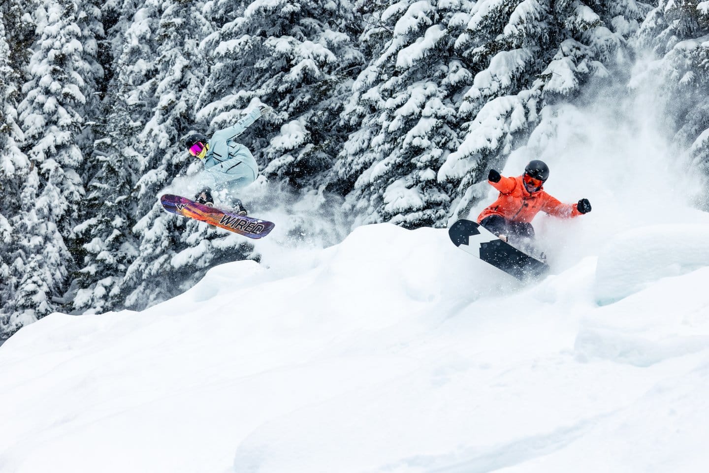Two snowboarders coming up and over a large pile of snow between lines of cedar trees. Their speed of approaching the jump has thrown up a lot of snow, creating a powdery background.