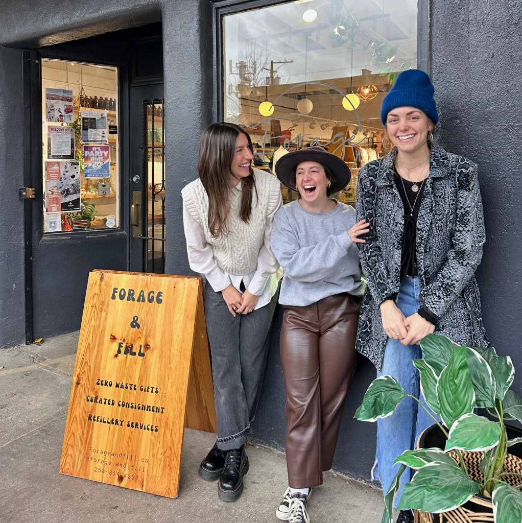 Three people standing in front of a grey stucco store front. A wood sign to the left of them reads Forage and Fill, a Revelstoke busniness