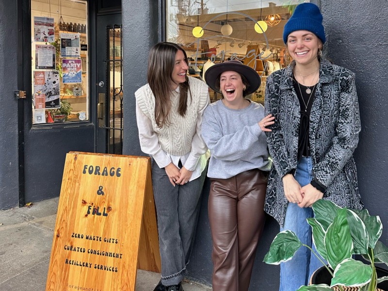 Three people standing in front of a grey stucco store front. A wood sign to the left of them reads Forage and Fill