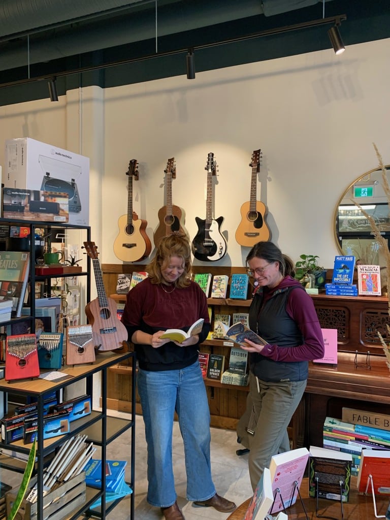 Revelstoke business Fable Book Parlour owners Stacy Batchelor and Kristin Olsen stand beside some shelves and look at open books in their hands.