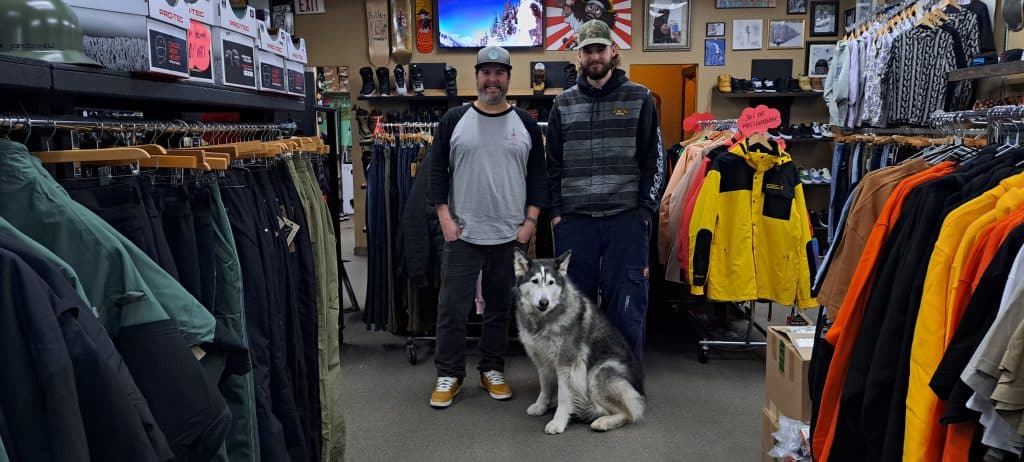 Two men stand in a shop surrounded by snowboard, ski and winter gear. In front of them sits a large husky cross dog who is staring off in the distance.