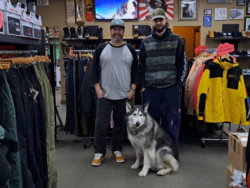 Two men stand in a shop surrounded by snowboard, ski and winter gear. In front of them sits a large husky cross dog who is staring off in the distance.