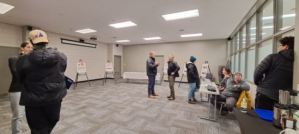Residents chat with staff and council members at the Revelstoke 2025 budget open meeting, people are standing around in a well lit general room, there are posters in the back with graphs on them.