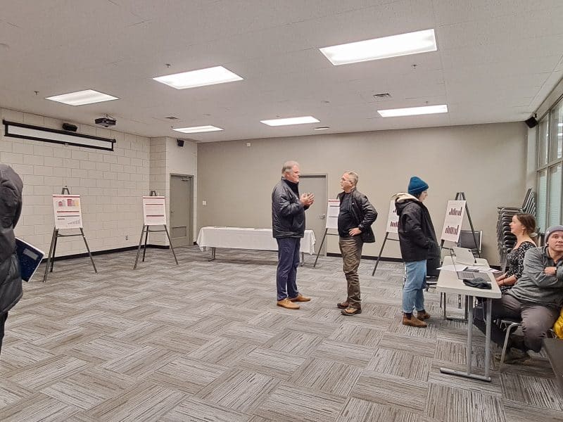 Residents chat with staff and council members at the Revelstoke 2025 budget open meeting, people are standing around in a well lit general room, there are posters in the back with graphs on them.