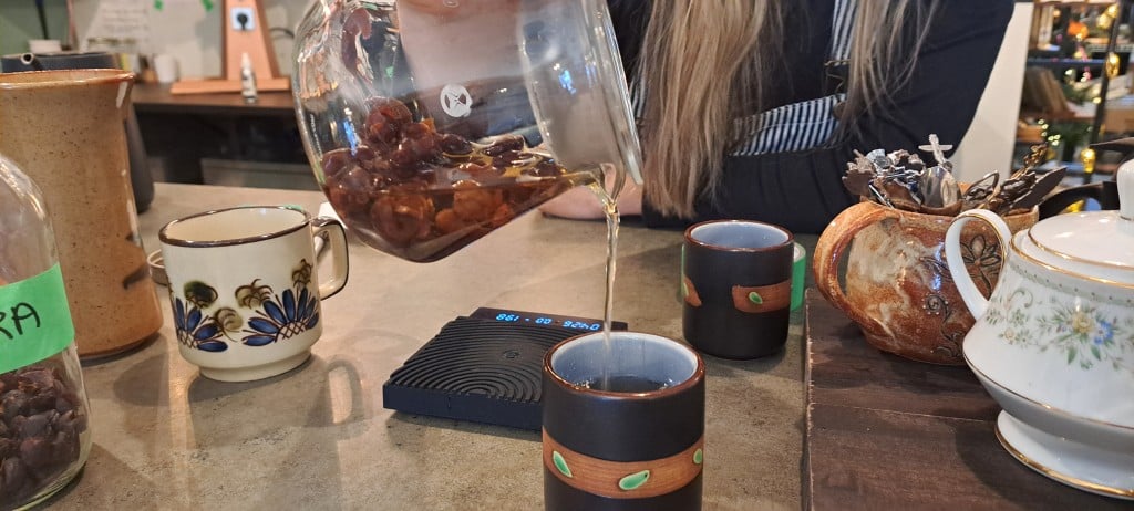 A glass infuser pours out a thin brown tea into a small mug. The diffuser is filled with dried berries