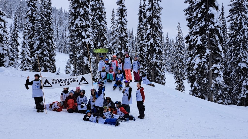 A collection of roughly 20 youth ages five to 12, all wearing various ski and snowboard gear, gather on one of the jumps. They are cheering at the camera, two are holding up a sign for Revelstoke snowboard club.
