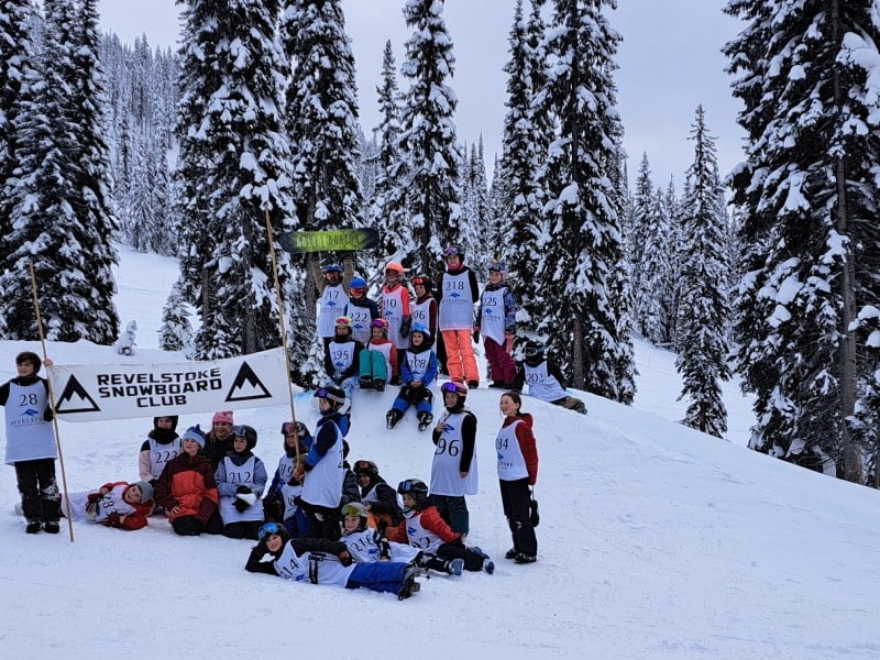 A collection of roughly 20 youth ages five to 12, all wearing various ski and snowboard gear, gather on one of the jumps. They are cheering at the camera, two are holding up a sign for Revelstoke snowboard club.
