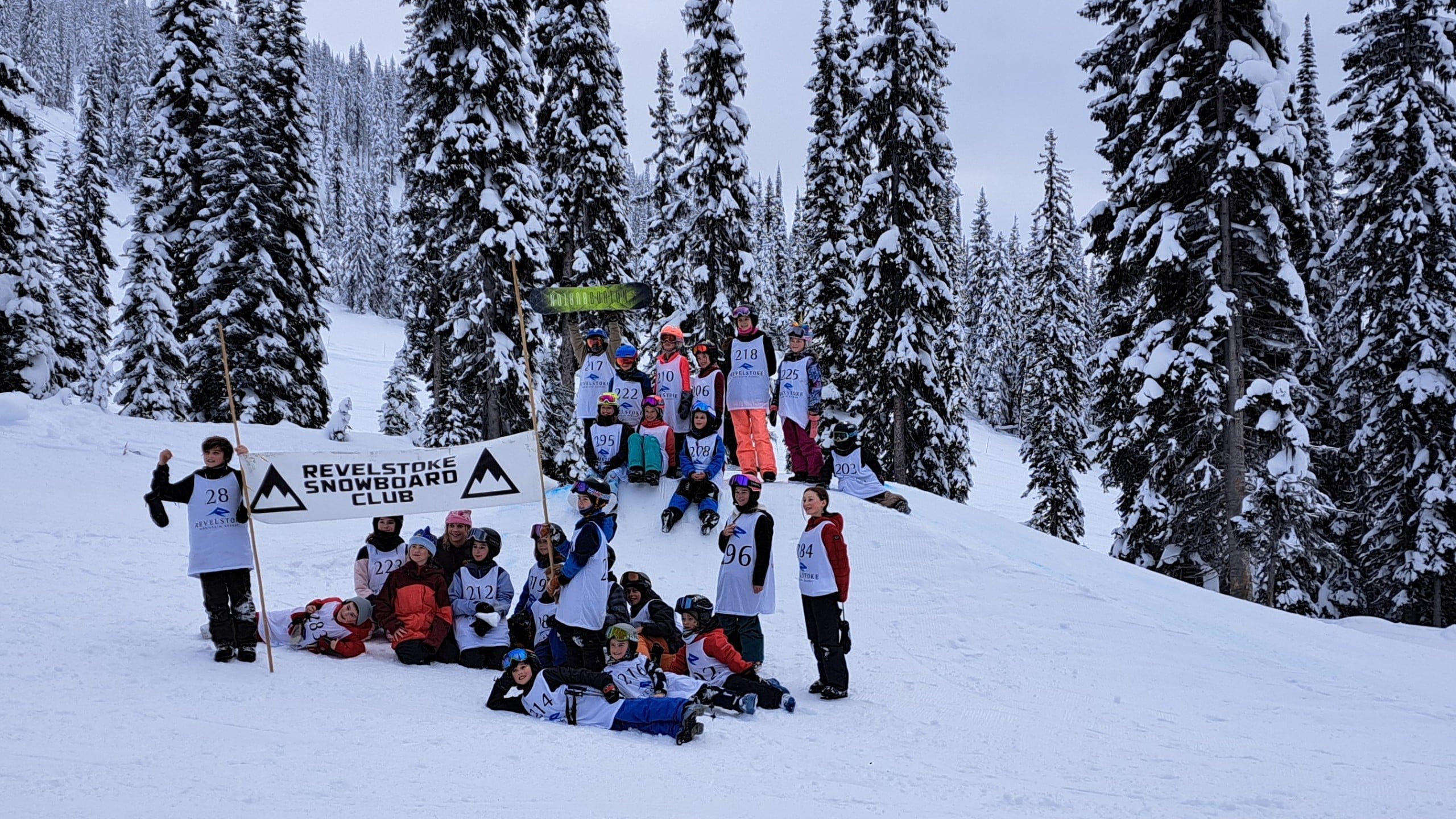 A collection of roughly 20 youth ages five to 12, all wearing various ski and snowboard gear, gather on one of the jumps. They are cheering at the camera, two are holding up a sign for Revelstoke snowboard club.