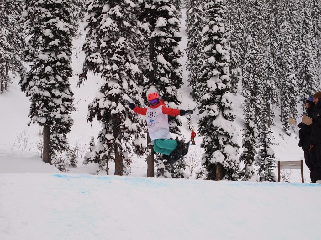 Emma Pfoster, a young snowboarder kicks their board sideways while coming off the kicker, a large mound of snow for jumping at Grom Con, Revelstoke Mountain Resort