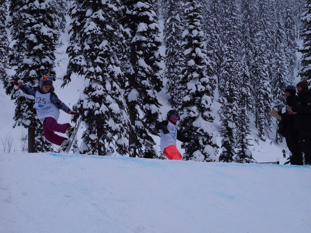 Two snowboarders clear the kicker, a large mound of snow for jumps, in tandem.