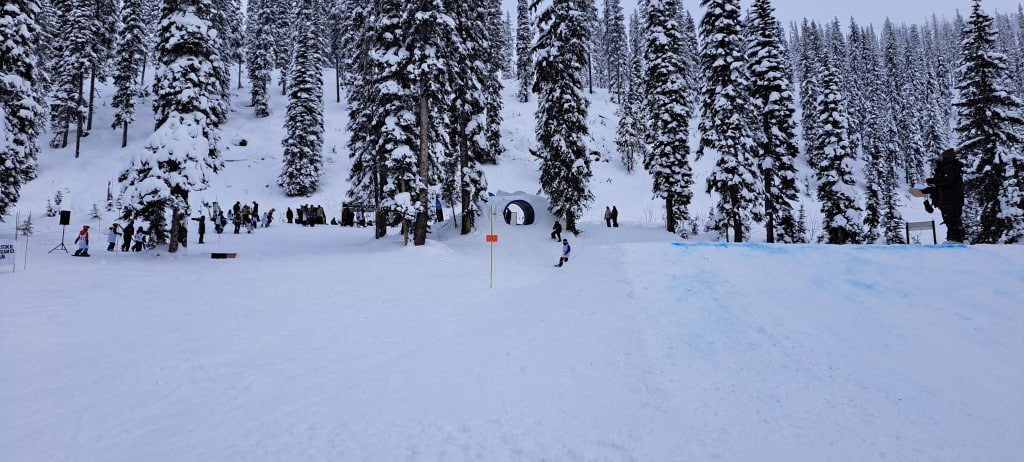Wide shot of the Gnome Zone terrain park with two rails, a box and a kicker. Grom Con youth participants line the top of the park readying for competition.