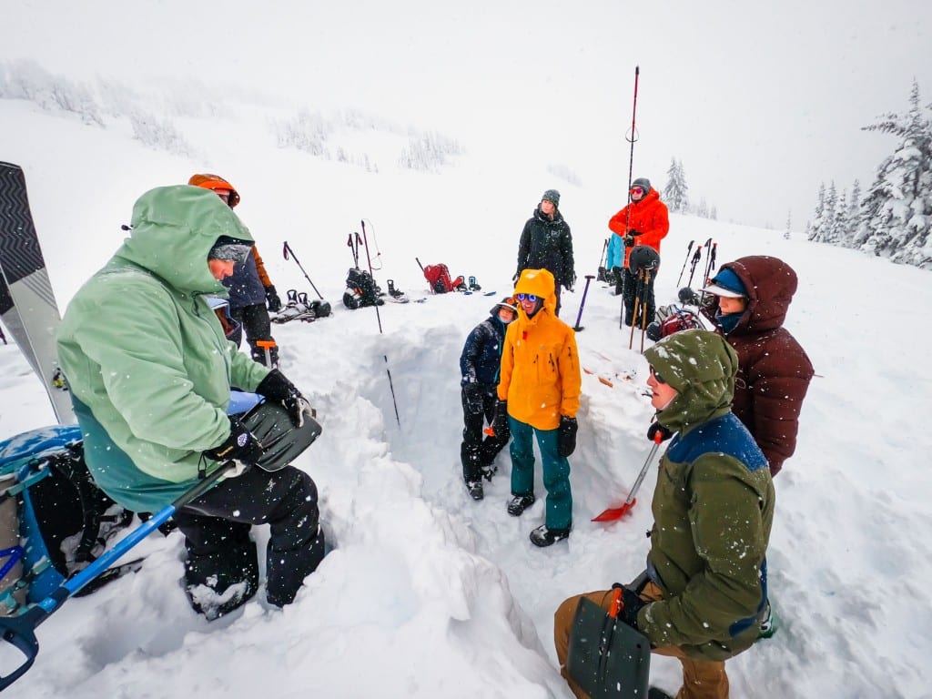 Open Mountains Project Society participants standing in a snowy valley watching an instructor show how to measure safe snow with a shovel in preparation of avalanche training.