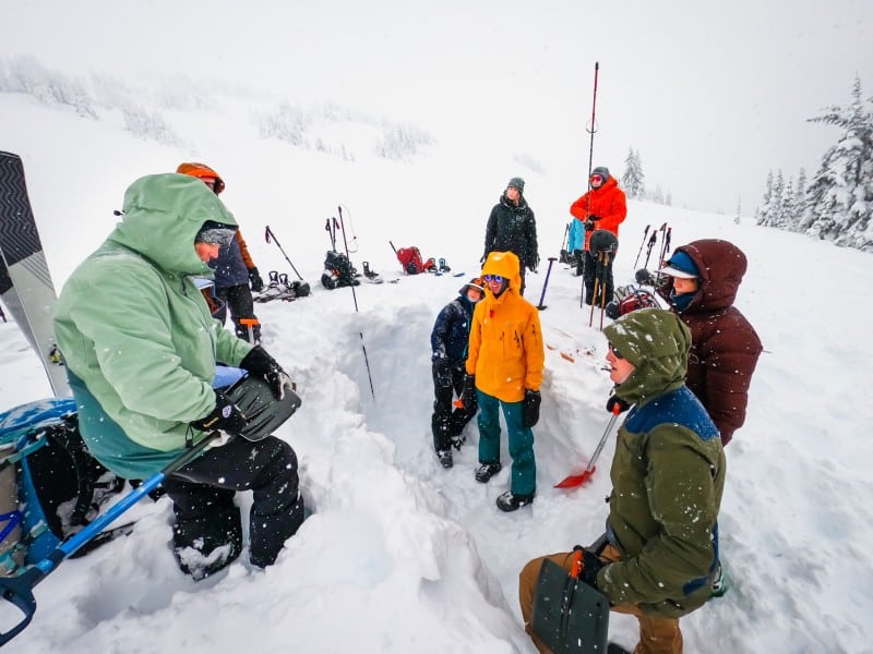 Open Mountains Project Society participants standing in a snowy valley watching an instructor show how to measure safe snow with a shovel in preparation of avalanche training.