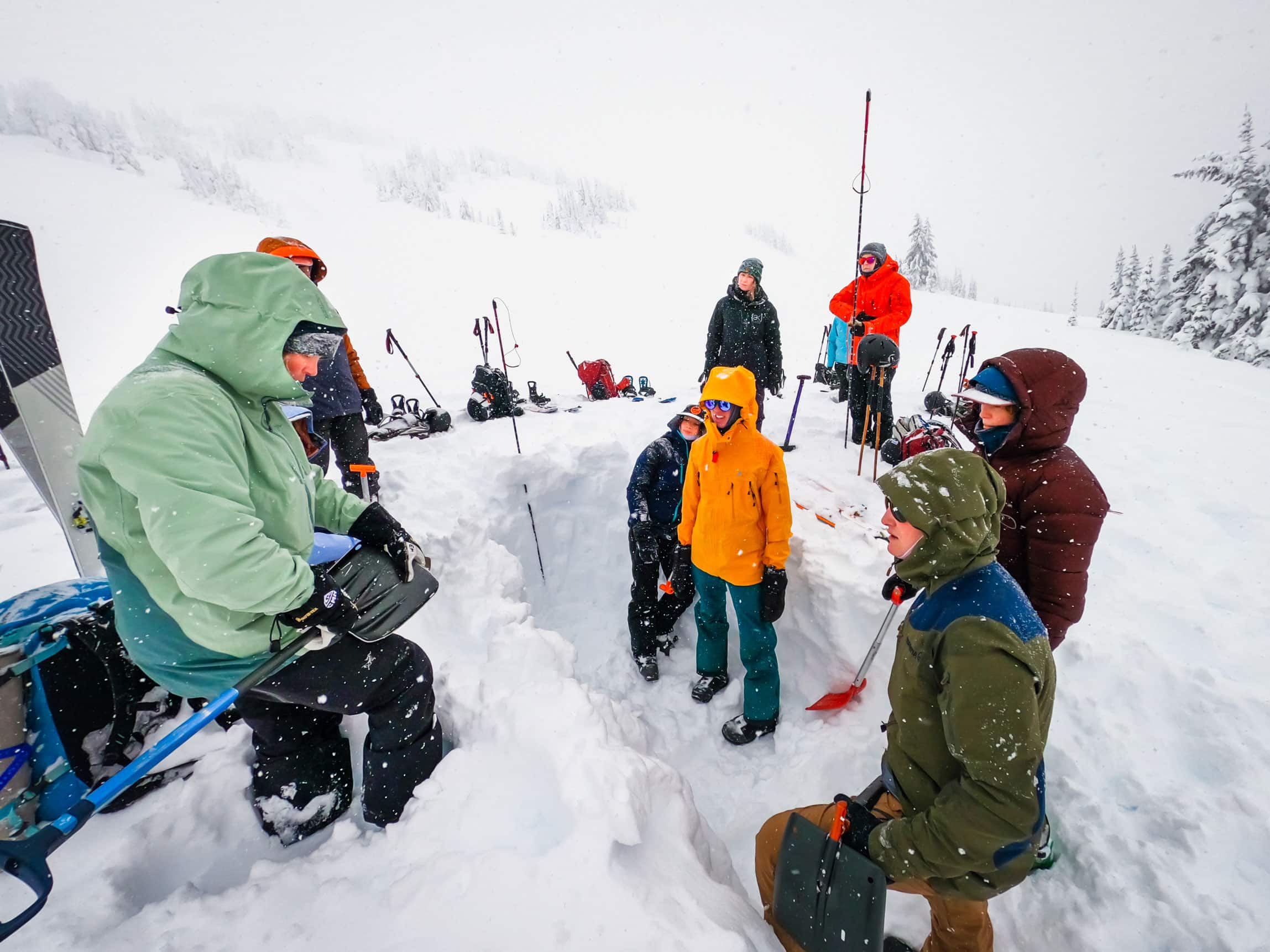 Open Mountains Project Society participants standing in a snowy valley watching an instructor show how to measure safe snow with a shovel in preparation of avalanche training.