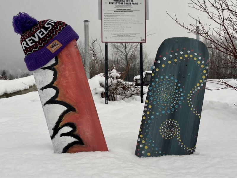 Painted skateboard decks without wheels or tracks, sticking out of a pile of snow. One is red with white mountains painted on it, one is green with dot patterns done in white. The red one is "wearing" a Revelstoke touque DonatedToHumanityInRevelstoke