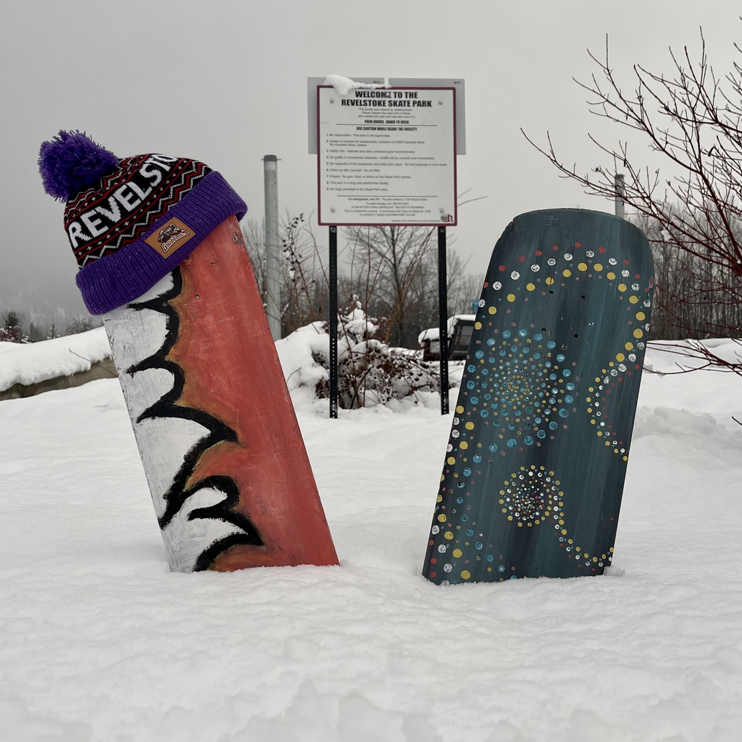 Painted skateboard decks without wheels or tracks, sticking out of a pile of snow. One is red with white mountains painted on it, one is green with dot patterns done in white. The red one is "wearing" a Revelstoke touque DonatedToHumanityInRevelstoke