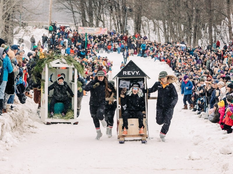 Two teams pushing large crate-like objects stylized as old wooden outhouses one skis. There is a teammate in either outhouse. Large crowds of people line either side of the track, standing on piles of snow.