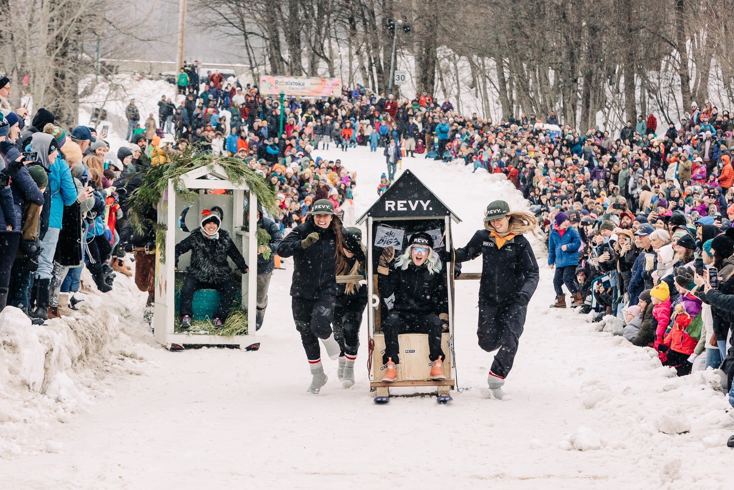 Two teams pushing large crate-like objects stylized as old wooden outhouses one skis. There is a teammate in either outhouse. Large crowds of people line either side of the track, standing on piles of snow.