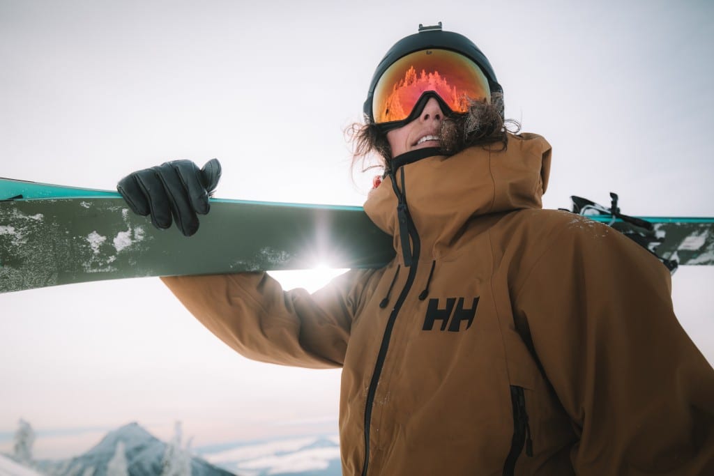 Nat Segal, an alpine skier wearing a helmet and goggles that obscures most of her face. She is holding a pair of skis over her right shoulder and looking over the camera. The sun is shining through the crook of her elbow. It is an action shot for the film Mountain Joy.