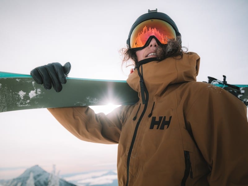 Nat Segal, an alpine skier wearing a helmet and goggles that obscures most of her face. She is holding a pair of skis over her right shoulder and looking over the camera. The sun is shining through the crook of her elbow.