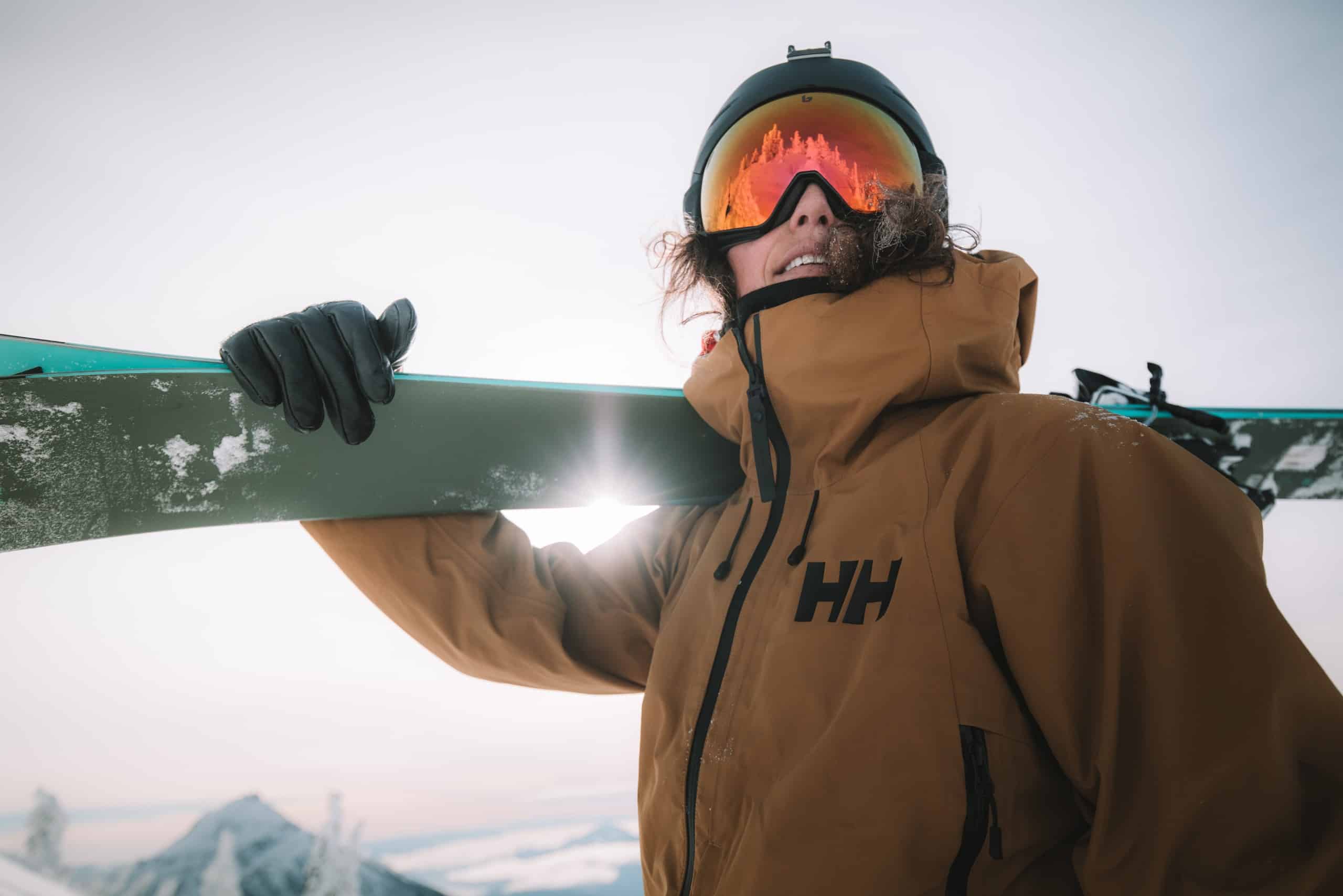 Nat Segal, an alpine skier wearing a helmet and goggles that obscures most of her face. She is holding a pair of skis over her right shoulder and looking over the camera. The sun is shining through the crook of her elbow.
