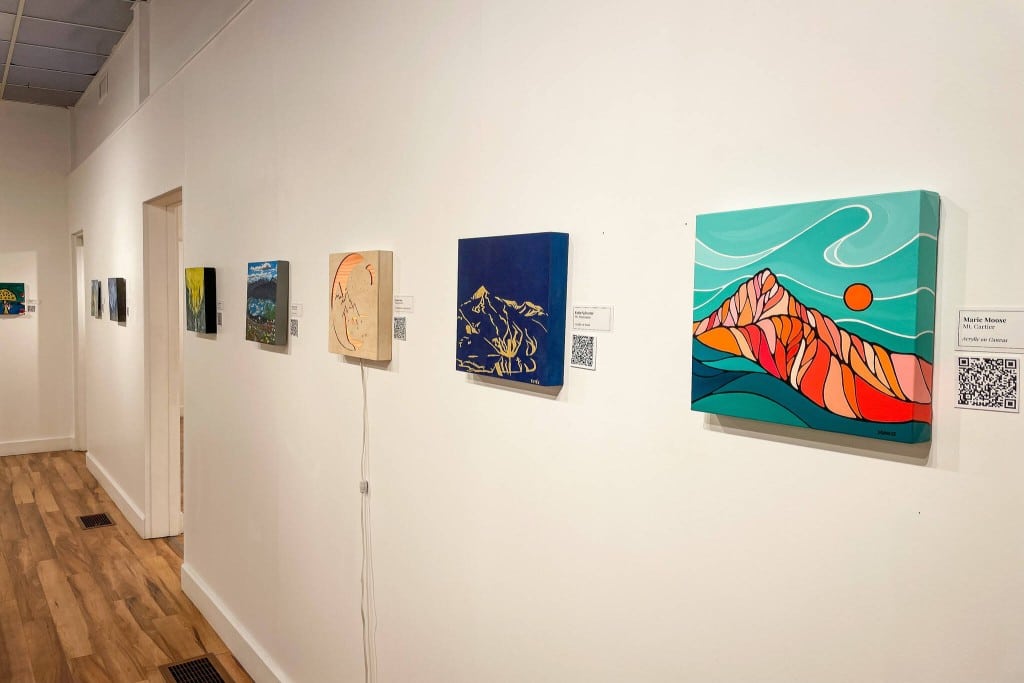 A large art gallery with a row of 12 inch by 12 inch pieces all hanging in a spaced out line roughly at eye level.