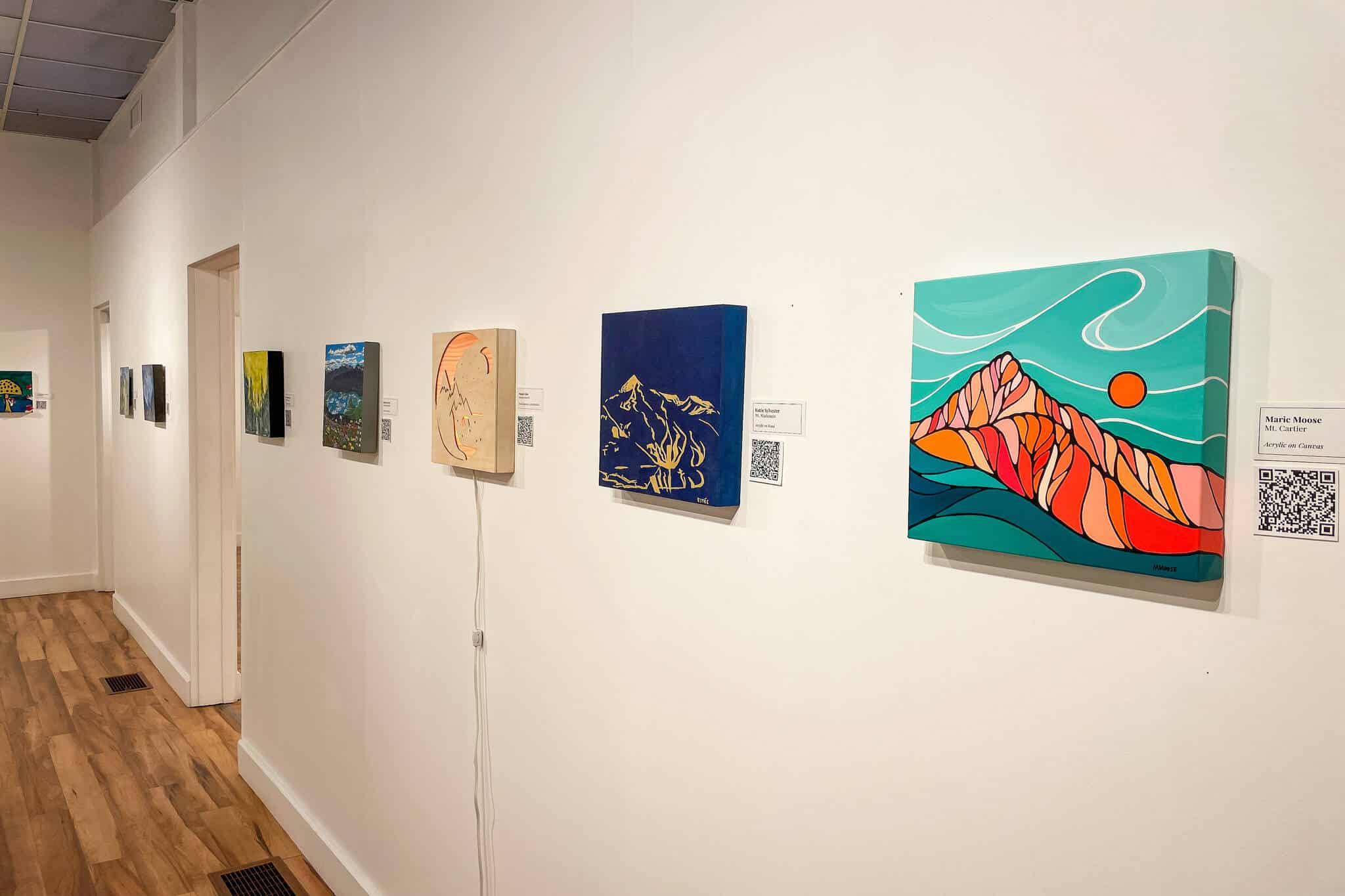 A large art gallery with a row of 12 inch by 12 inch pieces all hanging in a spaced out line roughly at eye level.