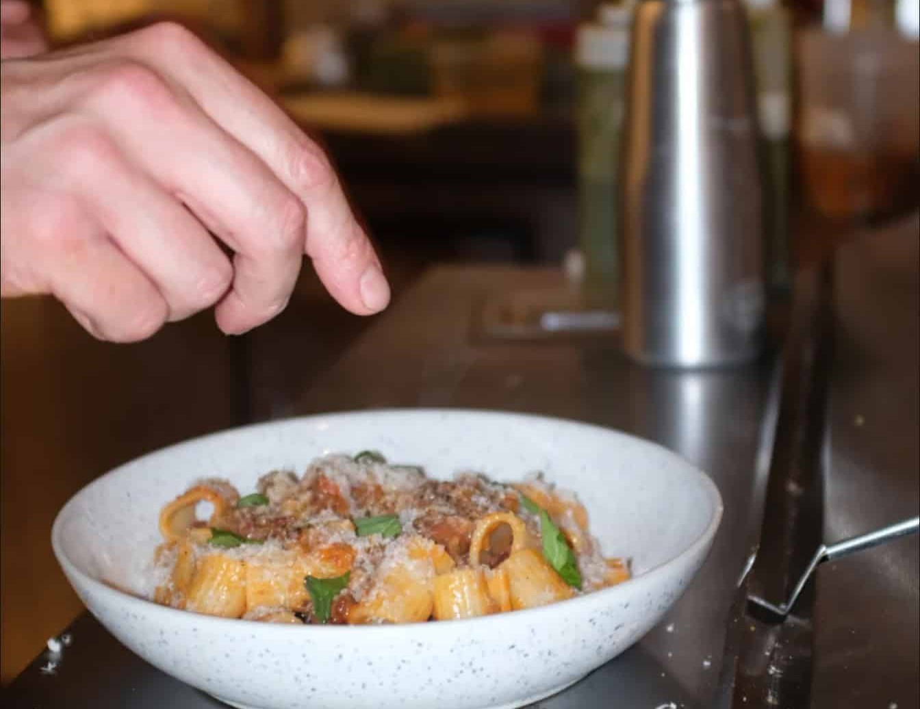 Close up shot of a pasta dish in a white bowl with a hand sprinkling garnishes over it at the Alfreds restaurant