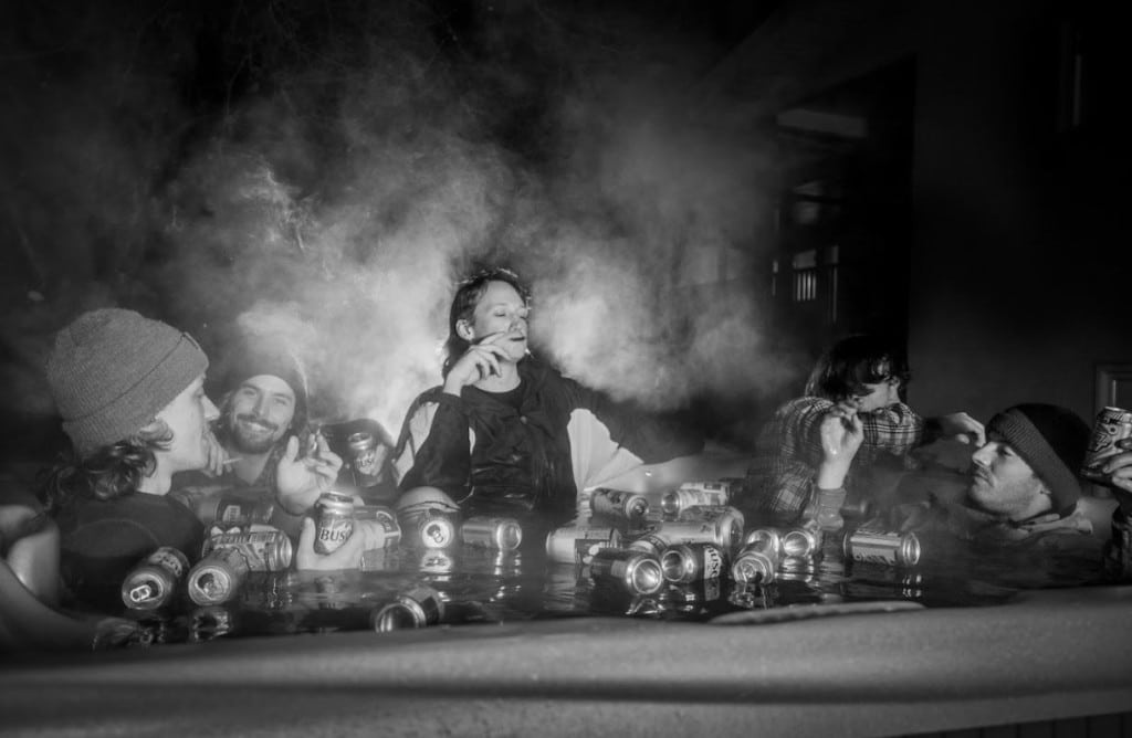 A black and white photo of the band Pickle Juice in a hot tub outdoors with a couple dozen empty beer cans floating on the water. There is a strong haze illuminated by a back light and one of the band members is standing in the hot tub, butting him slightly above the rest as he is illuminated by the back light. A couple of the members are smoking, everyone is wearing general layers of ski gear.