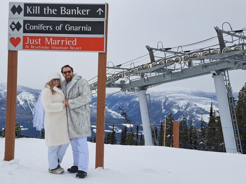 A couple dressed in fuzzy white jackets stand under a trail sign at Revelstoke Mountain Resort for the RMR Valentine’s Day weddings. One of the trail signs now says, "just married."