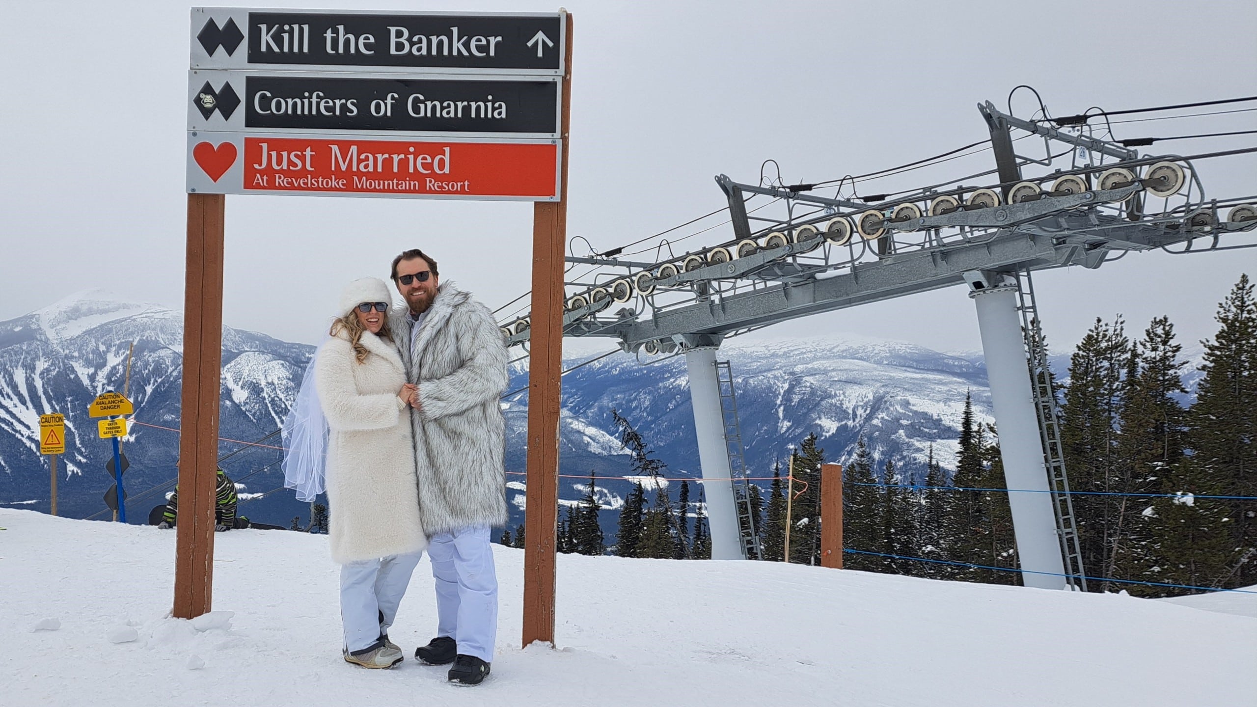 A couple dressed in fuzzy white jackets stand under a trail sign at Revelstoke Mountain Resort for the RMR Valentine’s Day weddings. One of the trail signs now says, "just married."
