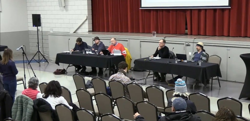 Revelstoke City Council members sit at two folding tables looking out onto a crowd of 20 participants asking questions to council in a large gymnasium setting.