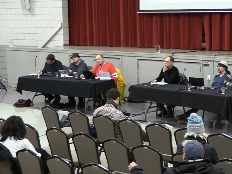 Revelstoke City Council members sit at two folding tables looking out onto a crowd of 20 participants asking questions to council in a large gymnasium setting.
