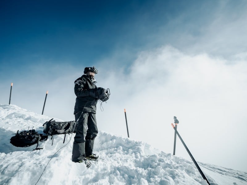 Gabriel Kocher, YETI Natural Selection drone pilot standing on the side of a run with his helmet goggles on and a large drone controller in his hands.