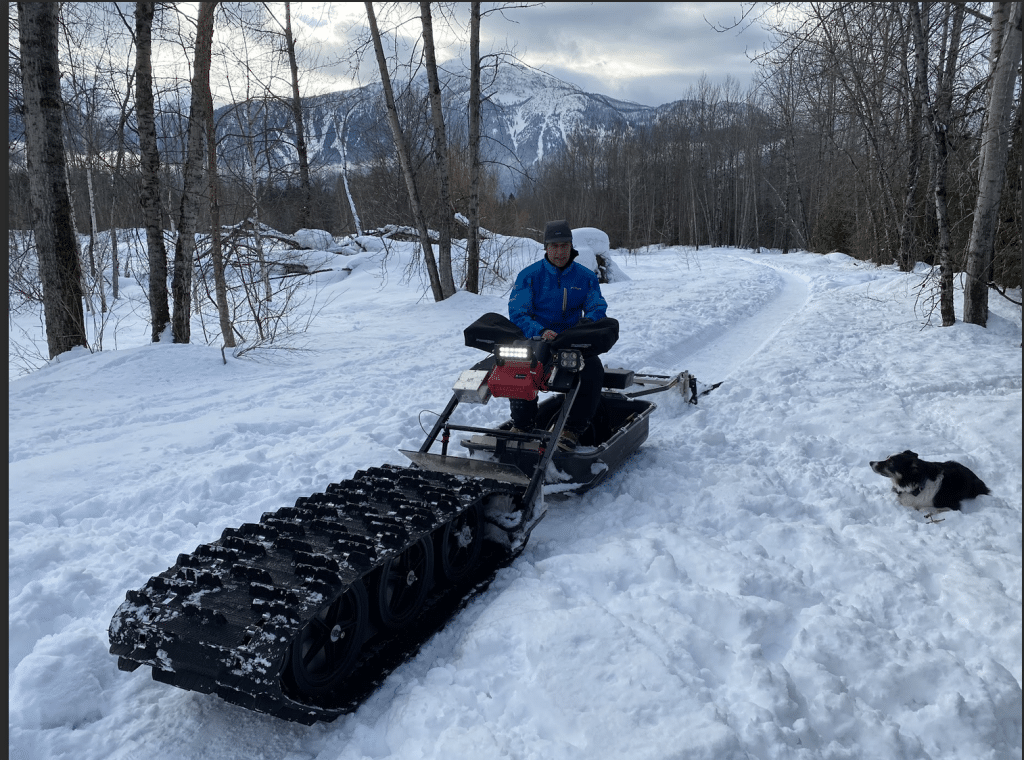 Ward Kemerer with Illecillewaet Greenbelt Society, dressed in a snow suit sitting on the modified track sled used to groom trails. The machine has a large groomer section on the front that juts out a couple of meters.