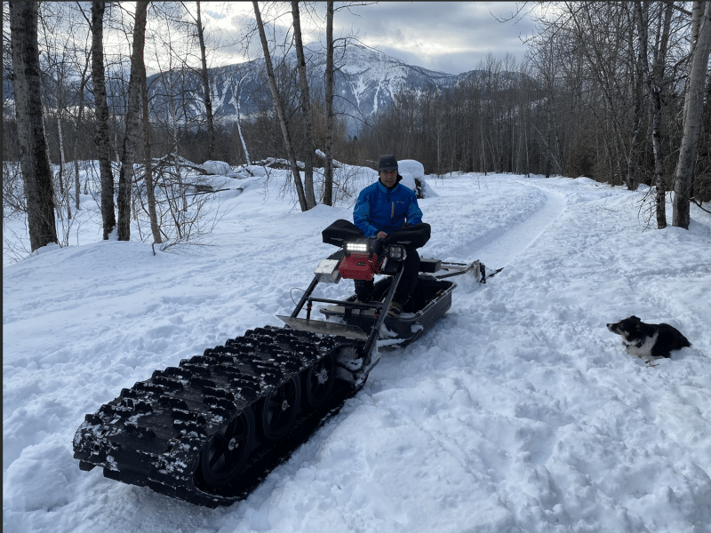 Ward Kemerer with Illecillewaet Greenbelt Society, dressed in a snow suit sitting on the modified track sled used to groom trails. The machine has a large groomer section on the front that juts out a couple of meters.