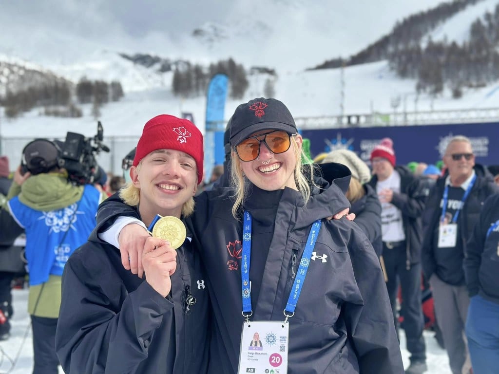 Yorke Parkin, left holding up his gold medal and smiling at the camera. Coach Saige Beaumont, right has her arm slung around his shoulder. Both are smiling widely at the camera. There is a collection of Special Olympics athletes and coaches behind them.