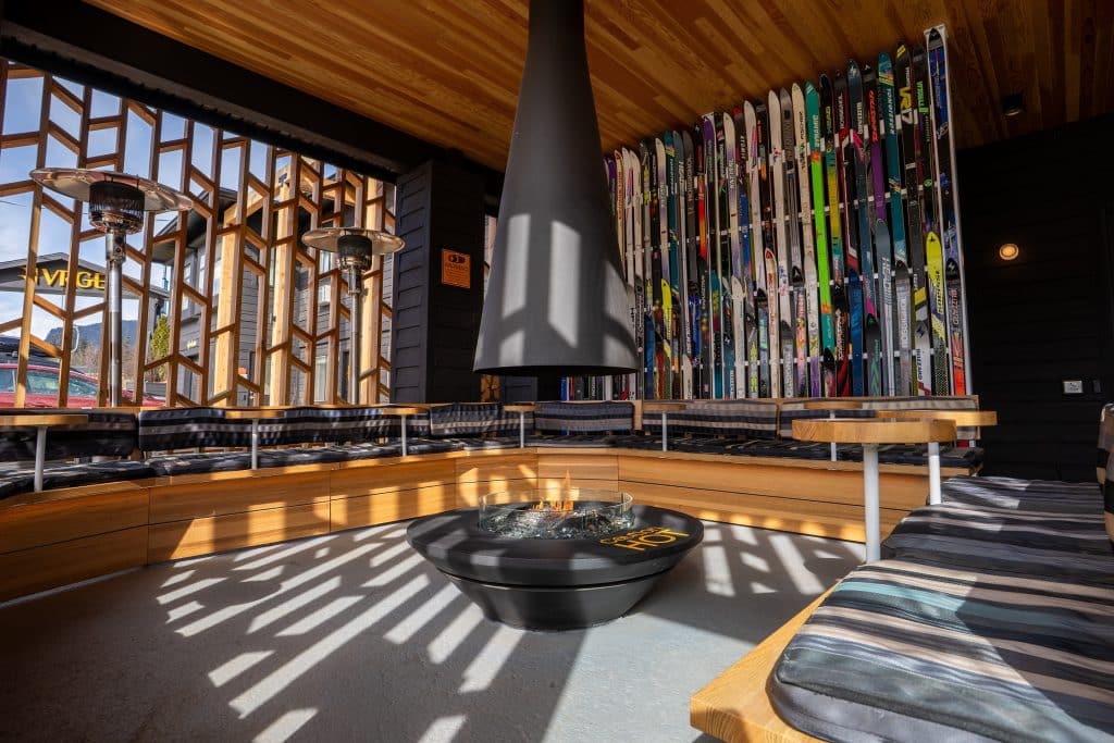 Shot of the VRGE Revelstoke hotel outdoor firepit loungue with wood lattice privacy walls on two sides and a chimney dropping from the centre of the roof over the glass fire bowl. Wood benches with outdoor cushions surround the firepit.