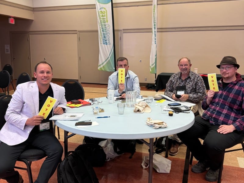 Revelstoke SILGA attendees councilor Matt Cherry, councilor Tim Stapenhurst, Mayor Gary Sulz and councilor Lee Devlin sit at a round table looking at the camera and all smiling in some capacity. They are each holding a yellow "Vote" card.