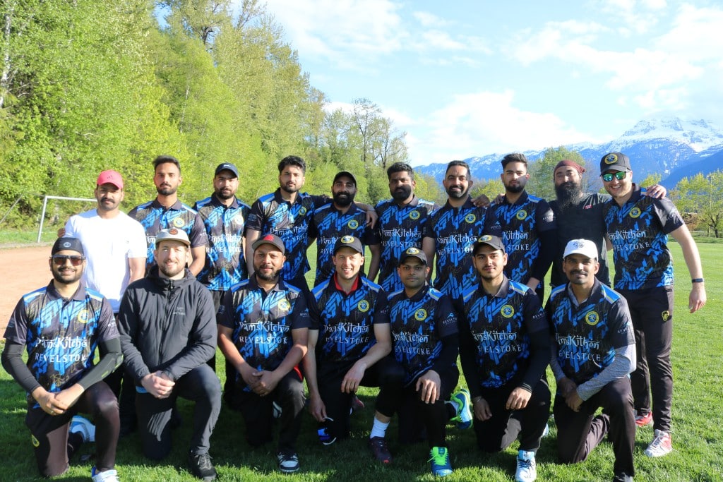 Revelstoke Cricket Club, 17 men of different ages and nationalities all wear the same black jersey with alpine blue stripes. Half of the team is standing in a line with arms over each others shoulders, the other half is in front of them taking a knee. All are smiling at the camera, they are outside on a bright blue sky day.