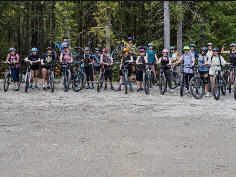 ShredHERS and ShredCOED participants on various mountain bikes line up side by side at the head of a trail. There is roughly 20 of them.