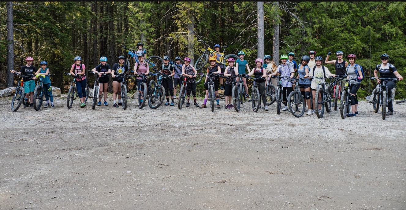 ShredHERS and ShredCOED participants on various mountain bikes line up side by side at the head of a trail. There is roughly 20 of them.