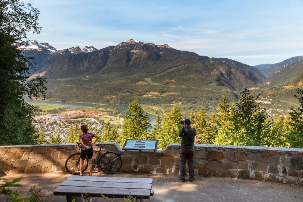 Two bikers stand on a large platform that gives a panorama view of the city of Revelstoke from Mount Revelstoke National Park